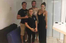 Four people are standing together in a lightly furnished room. Three are standing behind one, and they all appear to be smiling. Four people are standing together in a lightly furnished room. Three are standing behind one, and they all appear to be smiling.