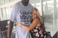 A man and a woman are posing together in a casual indoor setting. The woman is seated and hugging the man, who is standing and wearing a light blue T-shirt with the text "Savage by Nature. A man and a woman are posing together in a casual indoor setting. The woman is seated and hugging the man, who is standing and wearing a light blue T-shirt with the text "Savage by Nature.
