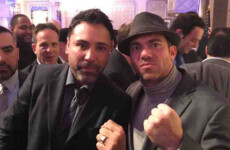 Two men pose together at an indoor event; one wears a suit and tie, and the other a hat and sweater, both making fist gestures. Two men pose together at an indoor event; one wears a suit and tie, and the other a hat and sweater, both making fist gestures.