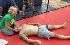 A shirtless man lies on a red mat with his eyes closed while a woman with short blonde hair attends to his head. A fenced area and other individuals are visible in the background. A shirtless man lies on a red mat with his eyes closed while a woman with short blonde hair attends to his head. A fenced area and other individuals are visible in the background.