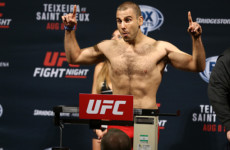 A shirtless man stands on a scale at a UFC event, pointing upwards with both hands and making a face, during a weigh-in. The background displays UFC branding and event details. A shirtless man stands on a scale at a UFC event, pointing upwards with both hands and making a face, during a weigh-in. The background displays UFC branding and event details.